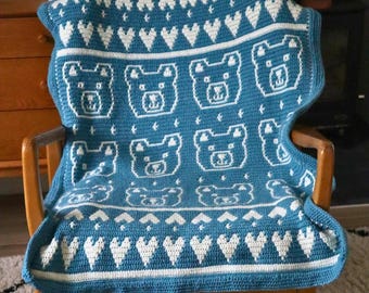 May include: A rectangular crochet blanket featuring a teal background with white patterns. The design includes rows of white hearts at the top and bottom, and a central section displaying repeating white bear face motifs in a grid layout.
