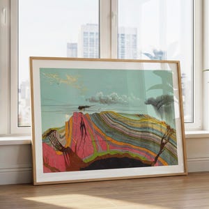 Puede incluir: Una impresión enmarcada que representa una sección transversal geológica de formaciones rocosas en capas. La obra de arte muestra una montaña con estratos rosas, naranjas y verdes bajo un cielo azul con nubes. La impresión está en un marco de madera clara, apoyada contra una ventana.