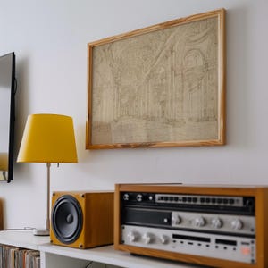 Op de afbeelding: Een ingelijste architectuurschets in een licht houten frame hangt boven een stereosysteem en een gele lamp. De schets toont een gedetailleerd interieur met bogen en kolommen. Het stereosysteem heeft een houten behuizing en zilveren componenten.