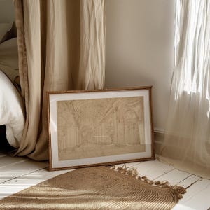 Op de afbeelding: Een ingelijste architectuurschets in een lichtbruine lijst leunt tegen een muur. De schets toont een gedetailleerd interieur met bogen en zuilen. Een geweven jute vloerkleed met kwasten ligt op de vloer en beige gordijnen omlijsten de sc&egrave;ne.