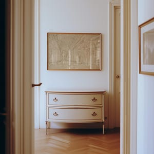 Op de afbeelding: Een cr&egrave;mekleurige dressoir met twee lades en gouden randen, tegen een witte muur. Boven de dressoir hangt een ingelijste architectonische tekening. De kamer heeft een parketvloer en is te zien door een open deur.