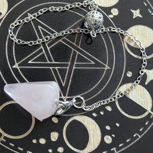May include: A rose quartz pendulum with a silver chain and a decorative silver ball at the top. The pendulum is a pale pink colour and is suspended over a black surface with white celestial designs.