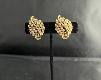 May include: A pair of vintage gold-tone clip-on earrings. Each earring features a cluster of small, cream-colored faux pearls within a gold-tone, leaf-shaped frame. The earrings are displayed on a black stand against a dark background.