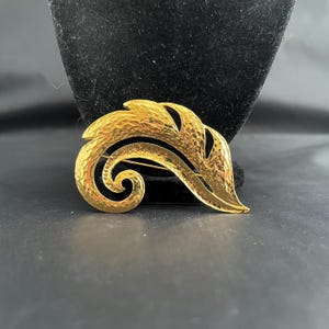 May include: A gold-toned brooch with a textured, swirling leaf design. The brooch has a pin backing for secure attachment. The design features a textured surface and a polished finish, creating a striking accessory.