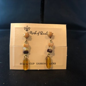 May include: A pair of gold tone screw-clip earrings with brown, beige, and amber coloured beads. The card reads "Mark of Quality Screw-Clip Earrings".