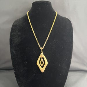 May include: A gold-toned necklace with a textured, diamond-shaped pendant. The pendant has an inner diamond shape and a small red accent. The necklace is displayed on a black mannequin.