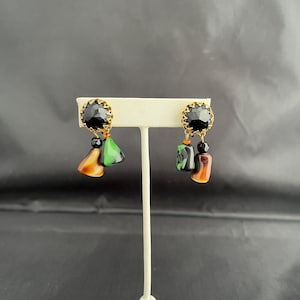 May include: A pair of gold-toned earrings with black, faceted center stones. Each earring features a cluster of colorful, dangling beads and geometric shapes in shades of green, brown, and black. The earrings are displayed on a white stand.