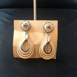 May include: A pair of white dangle earrings with silver accents. The earrings have a layered oval design with a small silver bead at the top.
