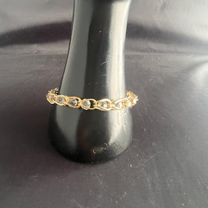 May include: A gold-toned bracelet with clear, faceted stones. The bracelet is displayed on a black, cylindrical object. The stones are set in a chain-like design, creating a delicate and elegant look. The background is a dark, solid color.