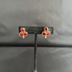 May include: A pair of gold-toned stud earrings, each featuring three round, vibrant red gemstones. The earrings have a delicate, leaf-like design, creating a floral or clover-like appearance. The gemstones are set in a gold metal setting.