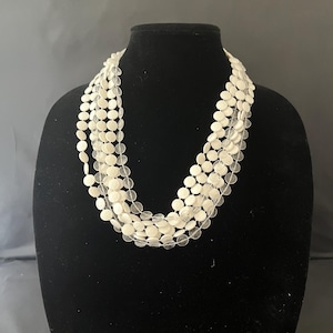 May include: A multi-strand necklace displayed on a black mannequin. The necklace features multiple strands of small, round, off-white beads. The beads are strung together, creating a layered effect. The necklace is a statement piece, suitable for various occasions.