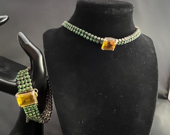 May include: A green beaded necklace and bracelet set. The necklace is displayed on a black mannequin head. Both pieces feature a large, square, amber-colored stone as a focal point. The bracelet is on a black hand-shaped display.