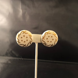 May include: A pair of white, round stud earrings with gold-colored filigree and small gold accents. The earrings are displayed on a white earring stand against a dark background. The earrings have a classic, vintage design.