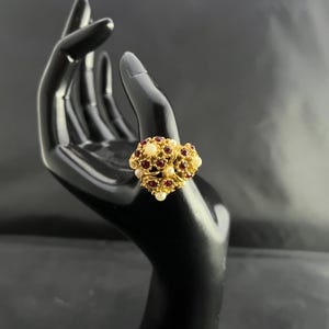 May include: A gold-toned ring featuring a cluster design. The ring is adorned with small, round, red gemstones and creamy white pearls. The ring is displayed on a black hand-shaped jewelry holder.