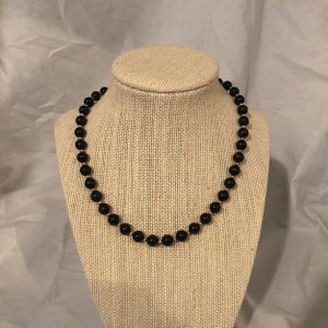 May include: A black beaded necklace with gold accents. The necklace is on a fabric-covered mannequin bust.
