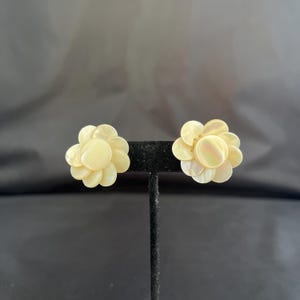May include: Two flower-shaped earrings with a pearlescent, off-white color. Each earring is composed of multiple petal-like pieces arranged in a circular pattern. The earrings are displayed on a black stand against a dark background.