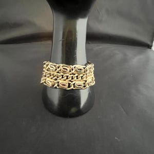 May include: A gold-toned bracelet with a chain link design. The bracelet is displayed on a black stand against a black background. The bracelet has three rows of interlocking links.