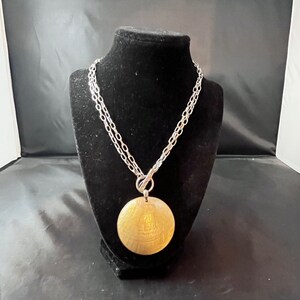 May include: A silver chain necklace with a large, round, gold-toned pendant. The pendant has a textured surface.