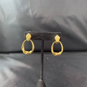 May include: A pair of gold-toned clip-on earrings with a twisted oval design. The earrings have a decorative detail at the top.