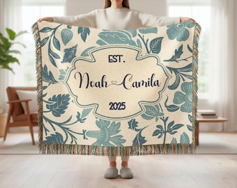 May include: A cream-colored throw blanket with a floral pattern in shades of blue and green. The blanket features a central design with the text "EST. Noah Camila 2025" in a decorative frame. The edges have a fringed trim.