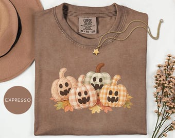 May include: A brown Comfort Colors t-shirt with a pumpkin graphic. The pumpkins have jack-o'-lantern faces and are in plaid patterns. A gold star necklace and a brown hat are also in the image. The shirt is size XL.
