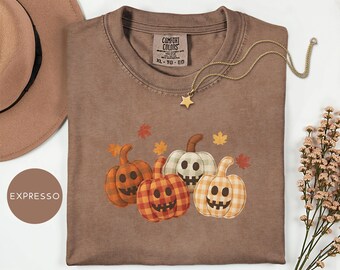 May include: A brown Comfort Colors t-shirt with a pumpkin graphic, featuring a plaid pattern and smiling faces. The shirt has a crew neck and short sleeves. A gold necklace with a star pendant is on the shirt. The shirt is size XL.