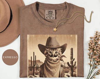 May include: A brown t-shirt with a graphic of a cat wearing a cowboy hat, mustache, and bandana. The cat is set against a desert landscape with cacti. A gold star necklace and a brown hat are also visible.