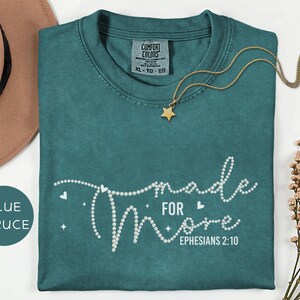 May include: Blue spruce t-shirt with the words "made for more" in white, along with the Bible verse Ephesians 2:10. A gold star pendant necklace and a brown hat are also in the image. The shirt is a Comfort Colors brand.