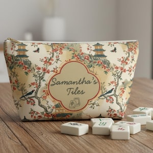 Op de afbeelding: Een beige etui met ritssluiting met een bloemen- en pagodepatroon, met de tekst "Samantha's Tiles". Verschillende witte mahjongtegels zijn gerangschikt op een houten oppervlak, als aanvulling op het thema van de etui.