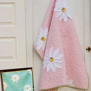 Könnte beinhalten: Eine rosafarbene Steppdecke mit weißen Gänseblümchen-Applikationen, die an einer weißen Tür hängt. Die Steppdecke hat einen grün-rosa floralen Rand. Eine kleinere grüne Steppdecke mit weißen Gänseblümchen-Applikationen hängt unter der größeren Steppdecke. Der Text "bloom" und "a quilt pattern by sara collett" ist über den Steppdecken gedruckt. Der Text "#101" ist unter der kleineren Steppdecke gedruckt.