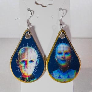 May include: A pair of teardrop-shaped earrings featuring a portrait of a character with pins in their head. The earrings have a dark blue background with a gold border and silver hooks. The image is a colorful, artistic representation.