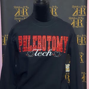 Könnte beinhalten: Schwarzes Langarmshirt mit rotem Glitzer-Schriftzug "PHLEBOTOMY Tech" mit zwei Herzen auf beiden Seiten des Wortes "Tech".