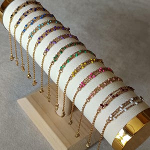 Könnte beinhalten: Präsentation von goldfarbenen Kettenarmbändern mit farbigen Perlenakzenten. Die Armbänder sind auf einem weißen, zylindrischen Ständer mit Holzsockel ausgestellt. Jedes Armband hat eine feine Kette und einen Anhänger.