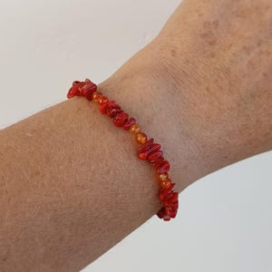 Peut inclure: Un bracelet composé de fragments de corail rouge et de perles orange. Le bracelet est enfilé sur un cordon souple et porté au poignet. Les éclats de corail sont de forme irrégulière, et les perles sont petites et rondes.