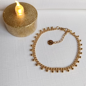 Peut inclure: Un bracelet doré orné de petites boules pendantes. Le bracelet est posé sur une surface blanche, à côté d'une bougie dorée scintillante avec une lumière jaune vacillante.