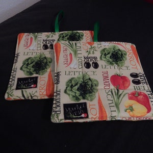 May include: Two beige pot holders with green fabric loops. The pot holders have a pattern of vegetables and text, including "Market Fresh", "Carrot", "Lettuce", "Tomato", "Scallions", "Olive", "Bell Pepper", and "Chili Pepper".