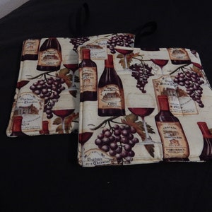 May include: Two wine-themed pot holders with a beige background and a pattern of wine bottles, grapes, and wine glasses. The text on the labels reads "Chateau de Chevaline" and "Chateau Saint-Estephe".