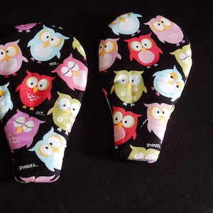 May include: Two black fabric oven mitts with a pattern of colorful owls. The owls have different expressions, some are sleeping and some are awake. The mitts have the word "snoozzl..." printed on them.