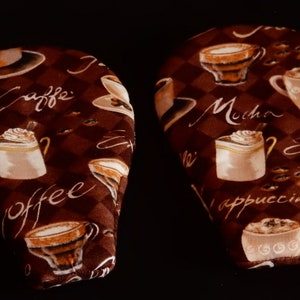 May include: Two brown fabric pot holders with a coffee theme. One pot holder has the words "Caffe" and "coffee" printed on it. The other pot holder has the words "Mocha", "Espresso", and "Cappuccino" printed on it.