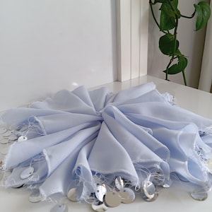 May include: A light blue, folded fabric with a circular design. The edges are adorned with silver sequins. The fabric is arranged in a radial pattern, creating a layered effect. The background is a white surface.