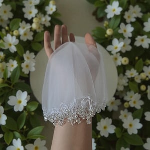 Puede incluir: Un velo de tul blanco con borde de cuentas. El velo se muestra sobre un fondo de flores blancas y hojas verdes. Accesorio nupcial.