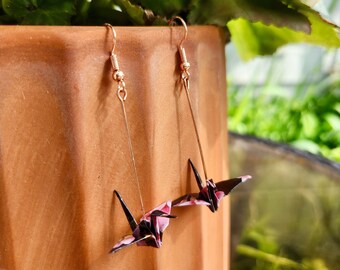 May include: A pair of gold earrings with pink and black origami cranes hanging from them. The earrings are hanging from a terracotta pot.