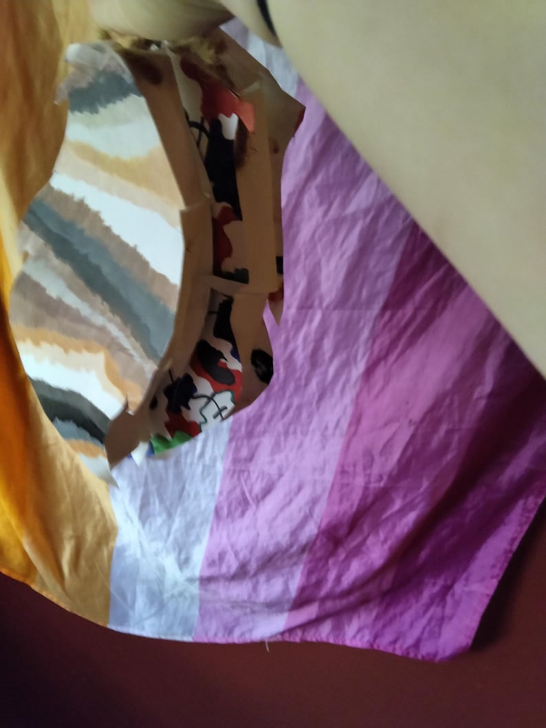May include: A folded fabric with a color block design. The fabric has a striped pattern in shades of yellow, white, gray, and brown. Another section of the fabric has vertical stripes in shades of pink and purple.
