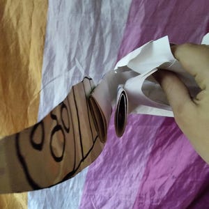 May include: A partially visible, light brown object with black outlines and circular designs, held against a backdrop of striped fabric in yellow, white, and purple. The object appears to be made of paper or cardboard.