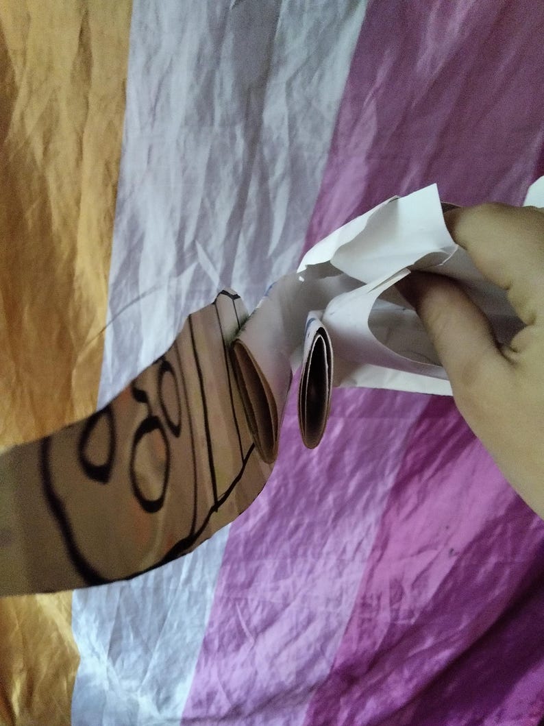 May include: A partially visible, light brown object with black outlines and circular designs, held against a backdrop of striped fabric in yellow, white, and purple. The object appears to be made of paper or cardboard.