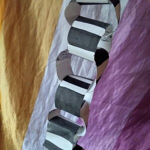 May include: A paper chain with alternating black and white stripes. The chain is made of folded paper and is hanging against a backdrop of yellow, white, and purple fabric.