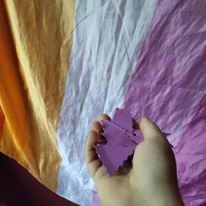 May include: A hand holding several small, purple, leaf-shaped cutouts against a backdrop of a draped flag in shades of orange, white, and purple. The flag's colors suggest a specific identity or cause.