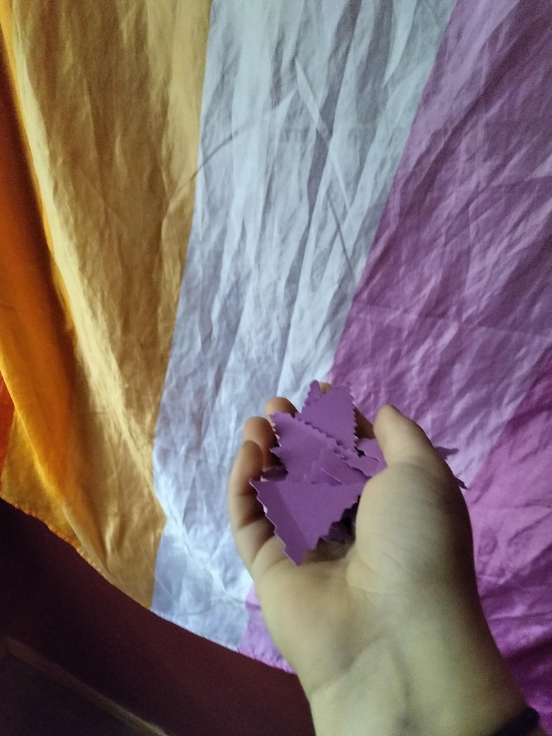 May include: A hand holding several small, purple, leaf-shaped cutouts against a backdrop of a draped flag in shades of orange, white, and purple. The flag's colors suggest a specific identity or cause.