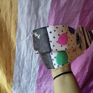 May include: A handmade paper mask with a black and white color scheme. The mask has a black section with the word "ROR" in white, and a white section with blue, pink, and green polka dots. The mask is held up by a person.
