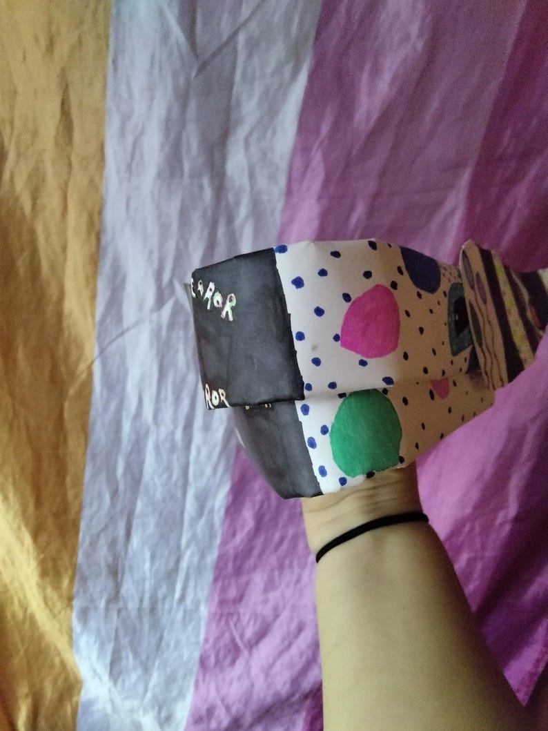 May include: A handmade paper mask with a black and white color scheme. The mask has a black section with the word "ROR" in white, and a white section with blue, pink, and green polka dots. The mask is held up by a person.